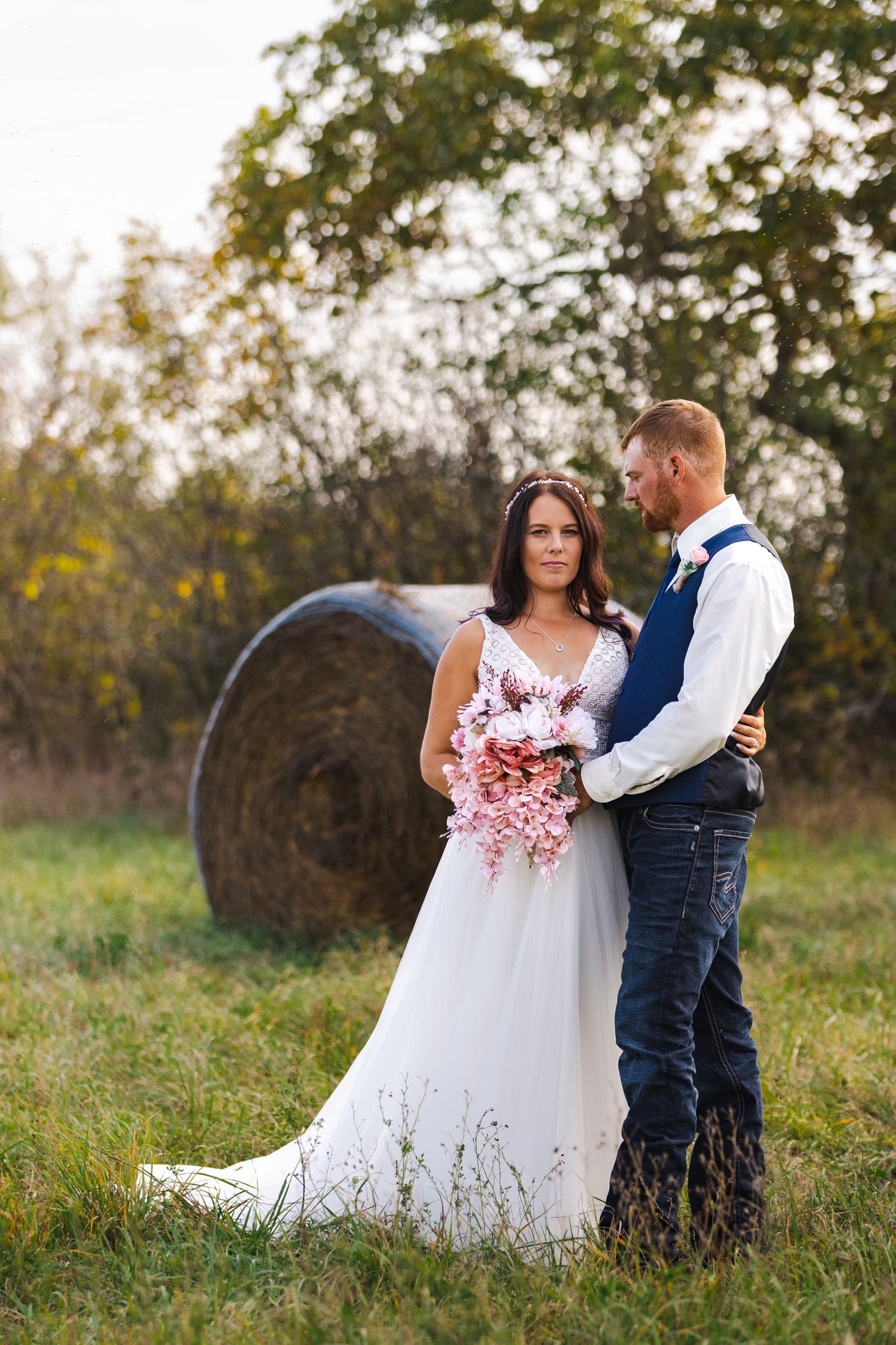 A couple in wedding attire embrace by a tree trunk near a serene lake on their special day.