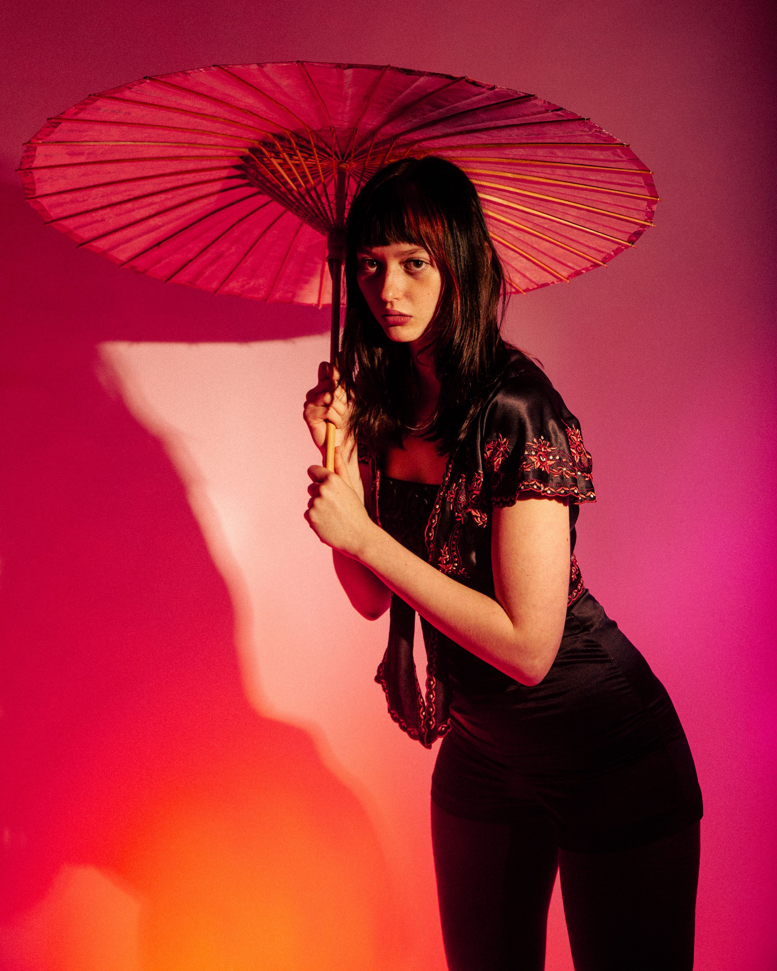 A figure in black poses with a red paper parasol against a pink background creating dramatic shadows.