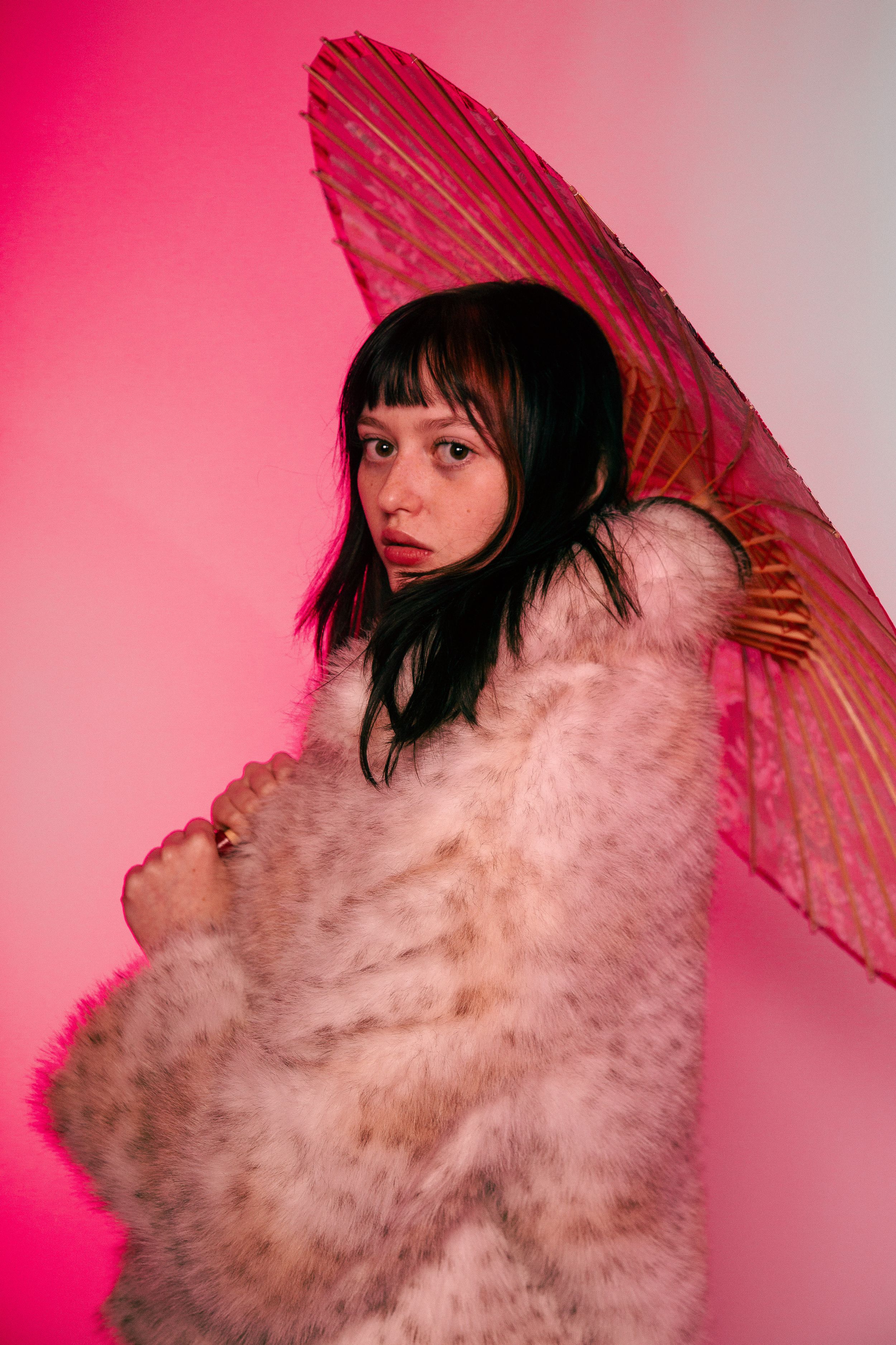 A figure in a fluffy white coat poses with a red parasol against a pink background in an artistic fashion series.