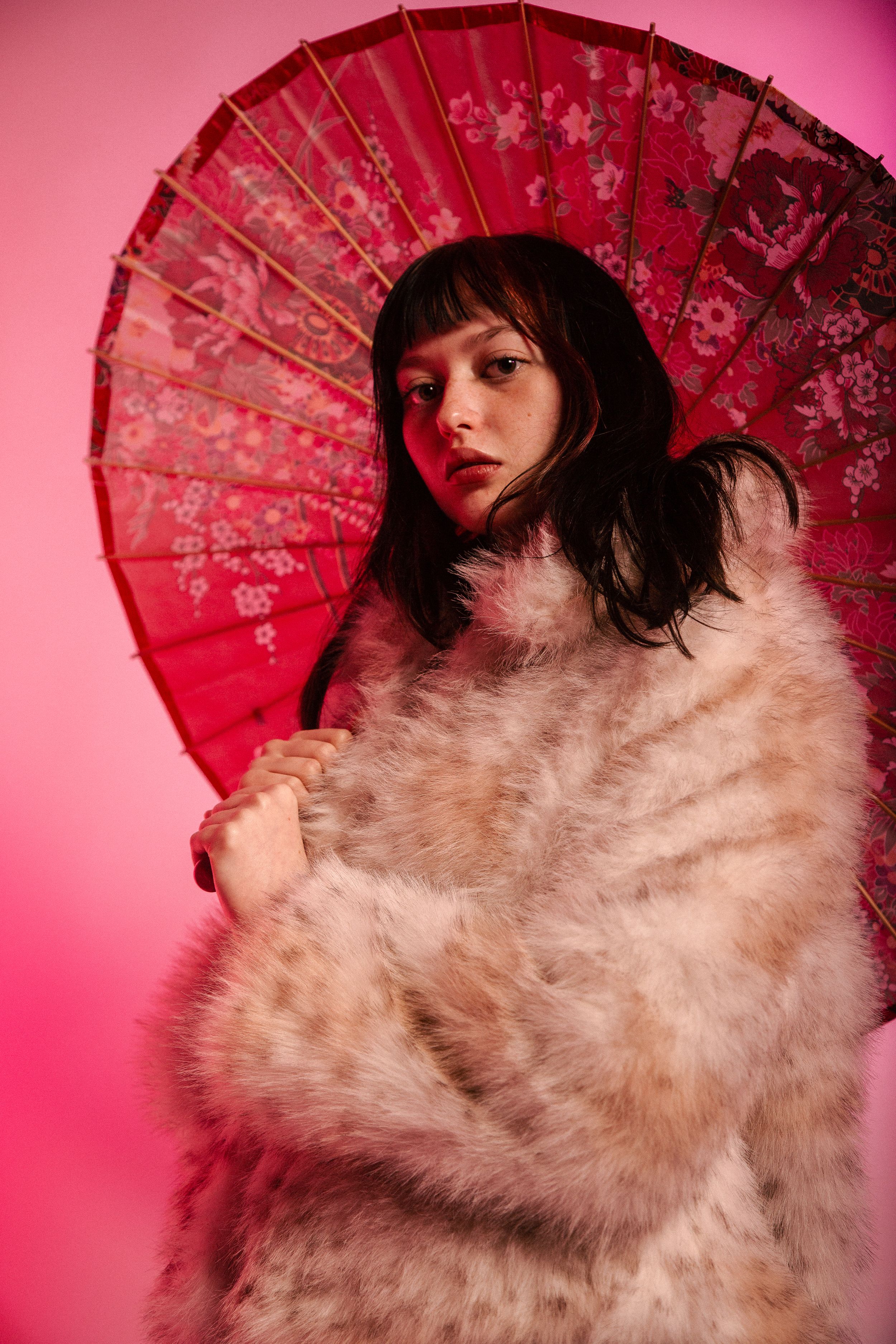 Dramatic fashion pose with beige fur wrap and red oriental umbrella against pink studio background.