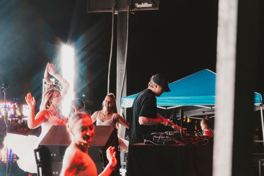 People dance and enjoy music at an outdoor nighttime event with DJ equipment and blue tent visible in background.