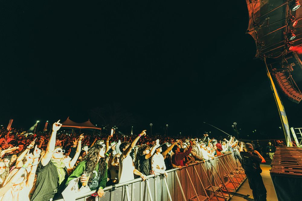 Concert crowd with raised hands at a nighttime outdoor music festival with bright stage lighting in the background.