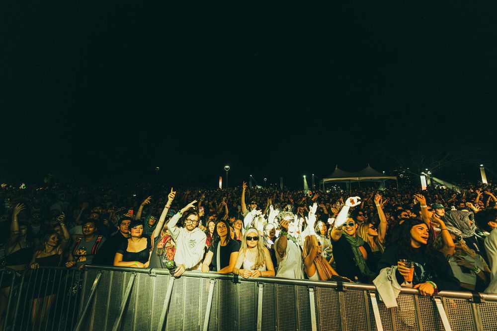 Concert crowd raises their hands in excitement during a nighttime performance at a music festival.