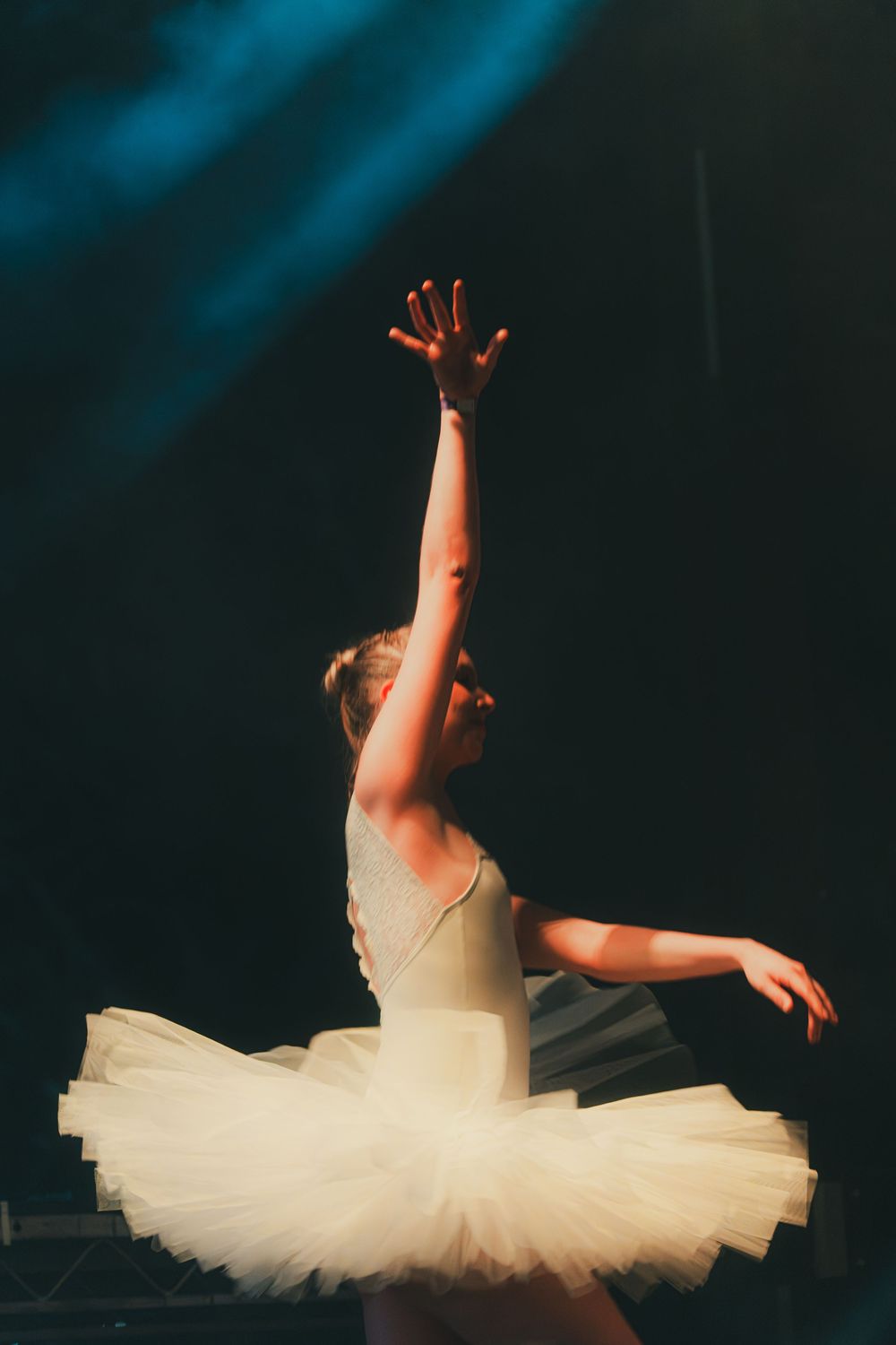 Ballet dancer in white tutu performs graceful movements on dark stage with dramatic lighting and ethereal atmosphere.