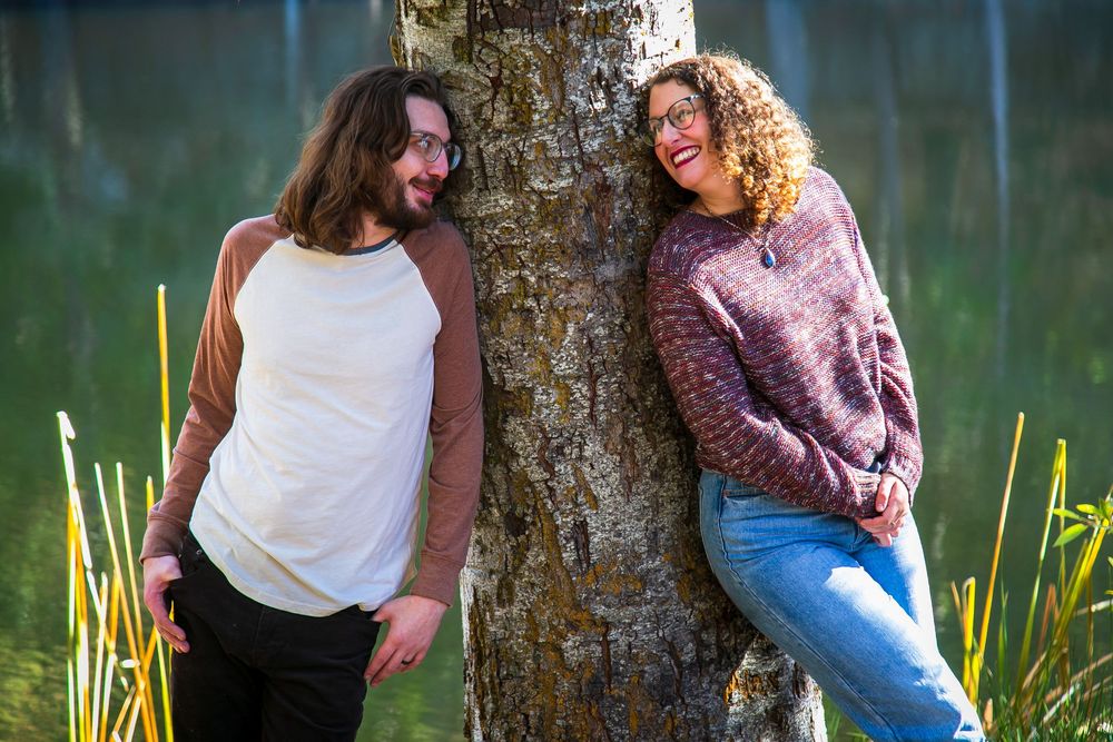 Two people leaning against tree trunk by lake with cattails in background.
