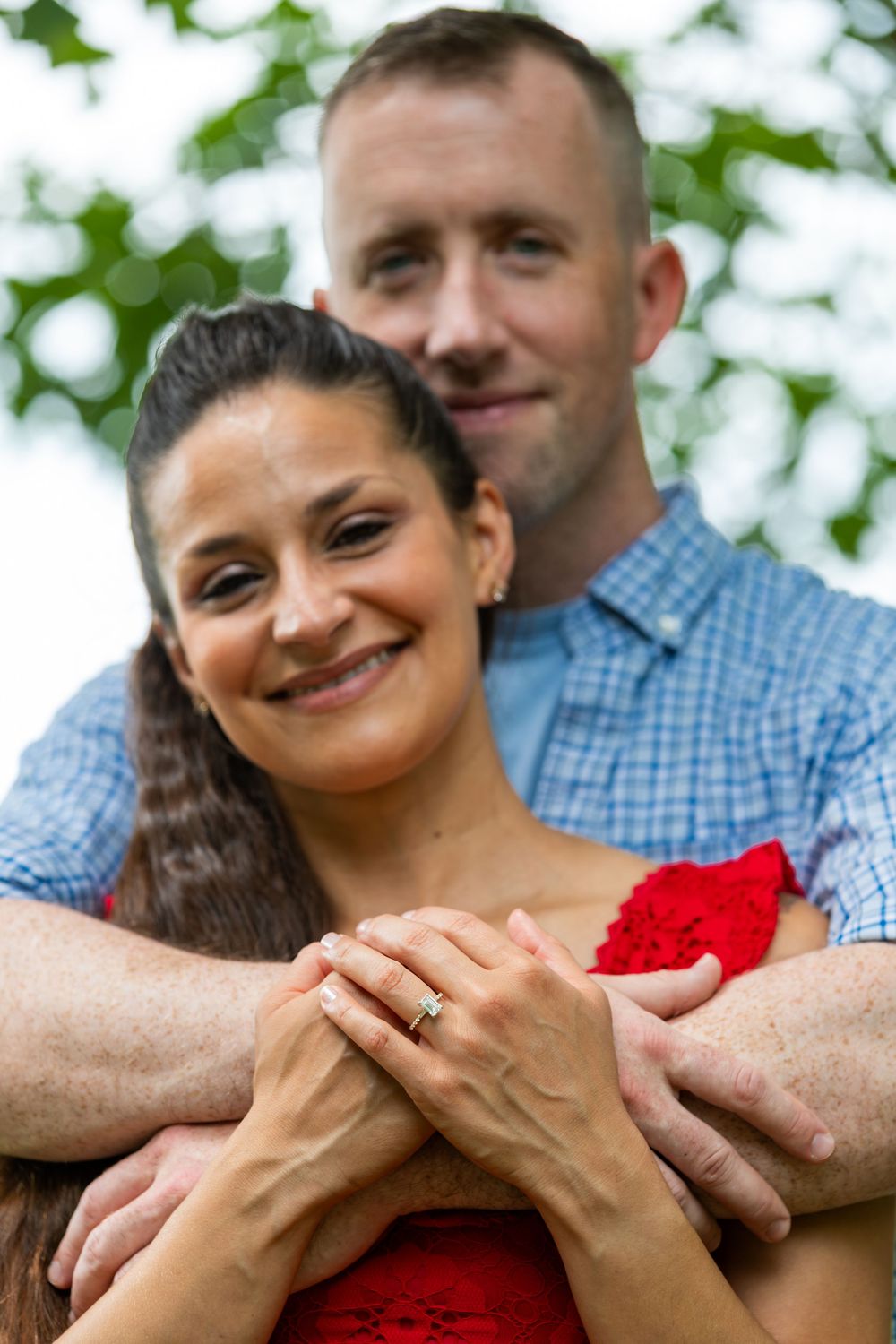 Smiling couple embracing outdoors, woman in red dress wearing engagement ring, man in blue shirt hugging her from behind.