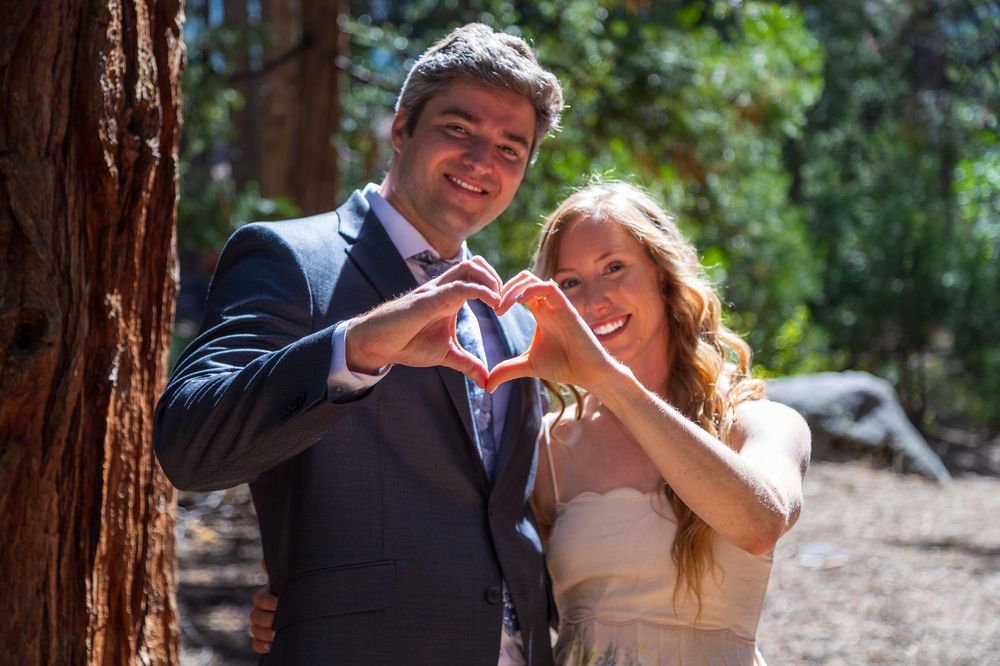 Couple making heart shape with hands against redwood tree in forest setting.