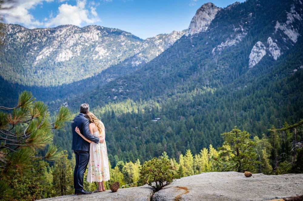 Couple embracing on mountain overlook with dramatic snow-capped peaks in the background.