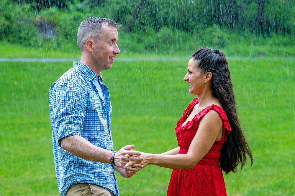 Couple holding hands and dancing in the rain on a lush green lawn, man in blue shirt and woman in red dress.