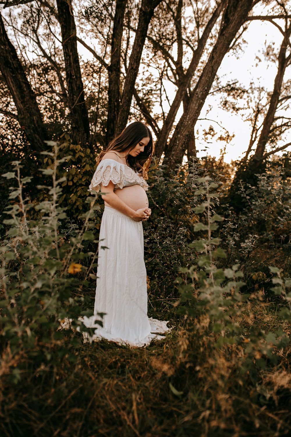 A ethereal maternity portrait in a flowing white dress among tall trees.