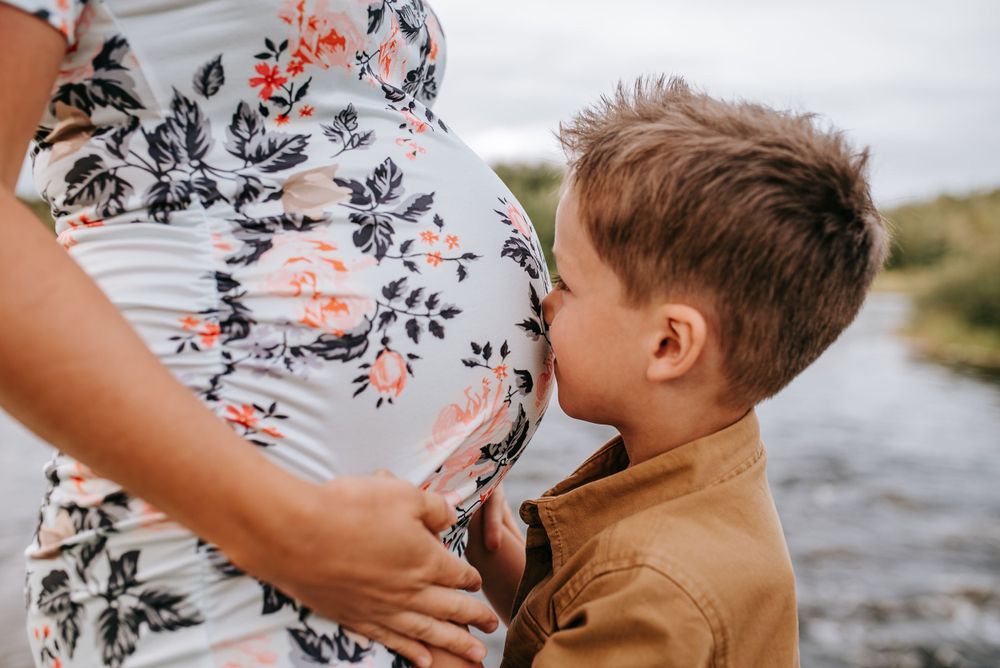 A child kisses a floral-dressed pregnant belly by a riverside.