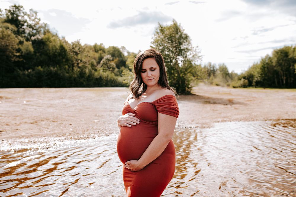 Beachside maternity photography with red flowing dress and dramatic cloudy sky backdrop.