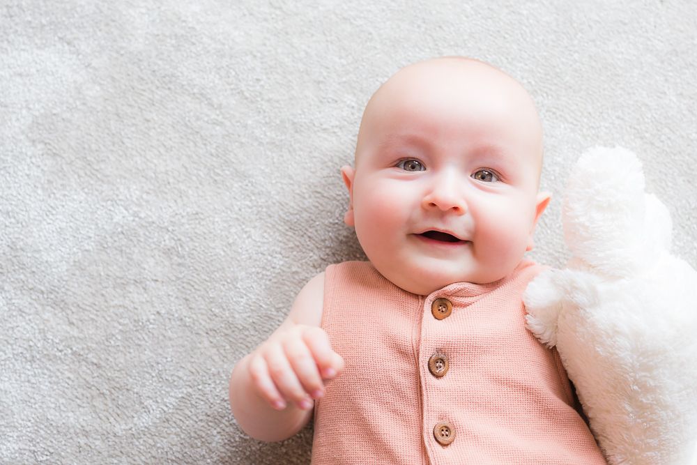 Baby wearing peach colored outfit smiling on soft grey blanket.