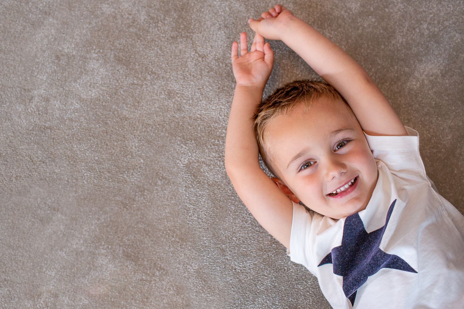 A joyful child in a tie lies on gray carpet with arms raised playfully.
