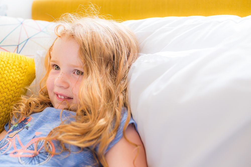 A child with long hair rests on white bedding next to a yellow pillow.