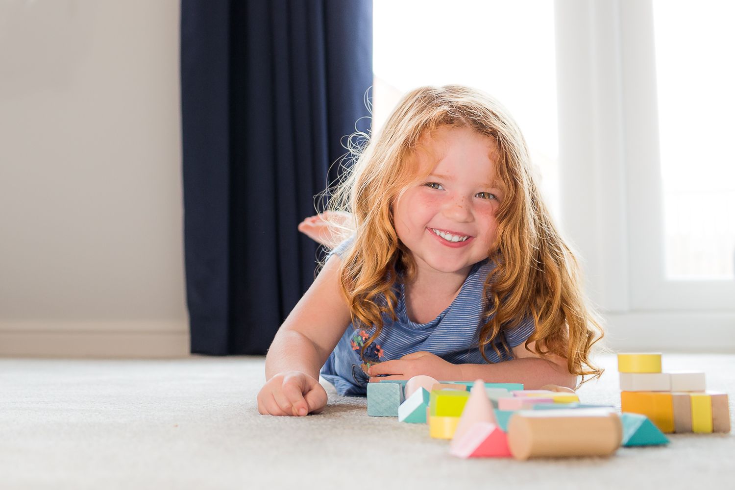 Child playing with colorful wooden building blocks on carpet next to blue curtains.