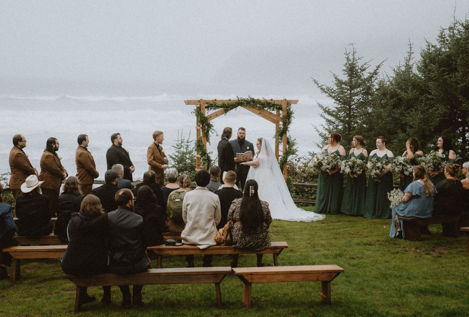 Outdoor wedding ceremony with couple at wooden arch, bridesmaids in green, ocean backdrop, guests on wooden benches.