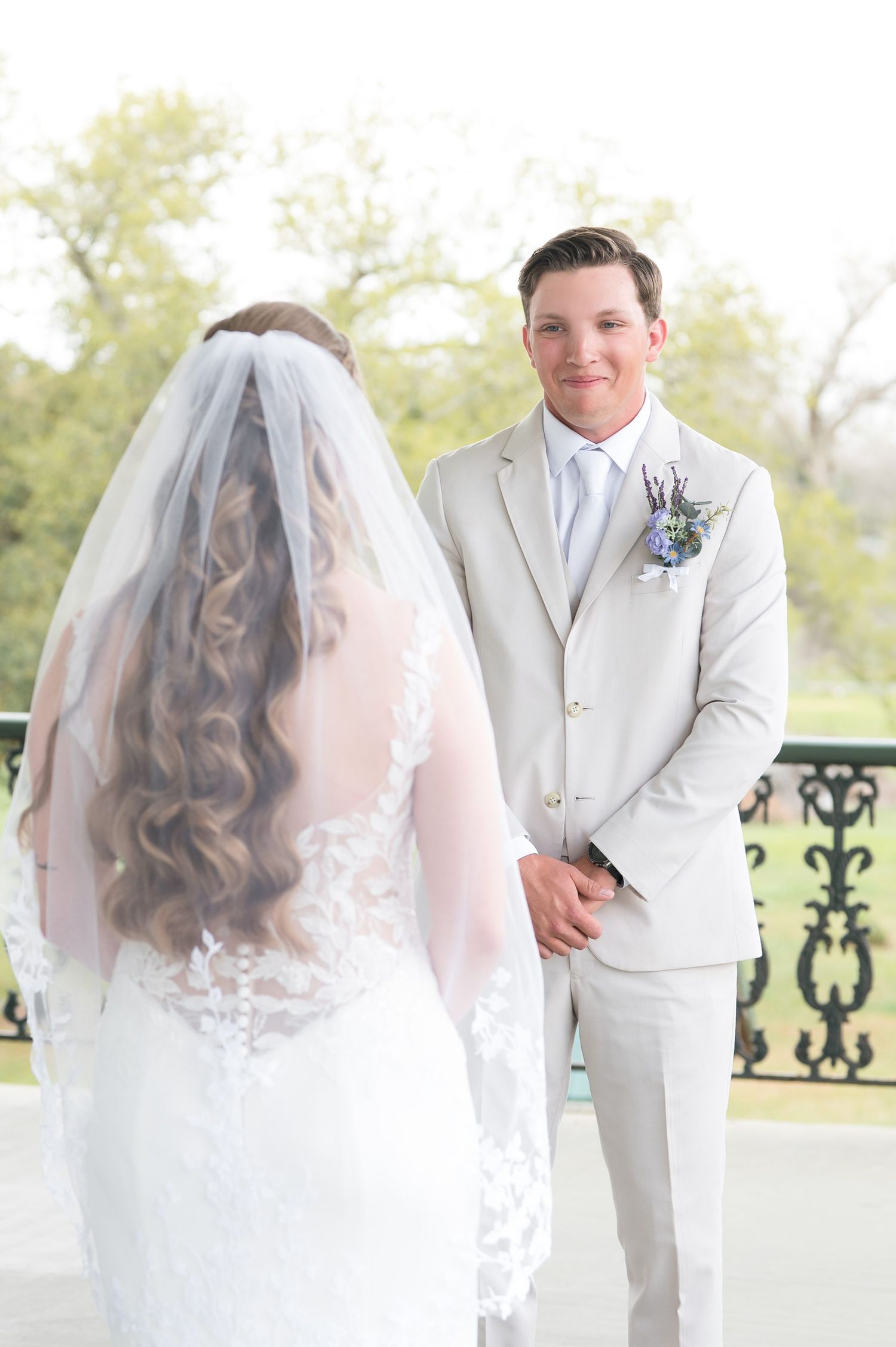 A bride in a white lace gown with long veil faces a groom wearing a light gray suit during their first look.