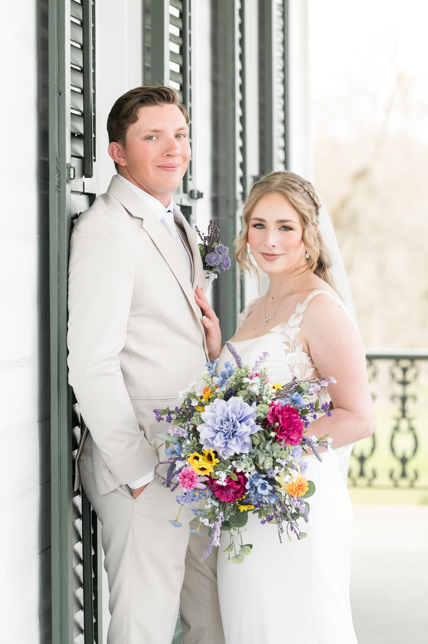 A wedding couple poses together on a porch with colorful spring bouquets featuring purple, pink and yellow flowers.