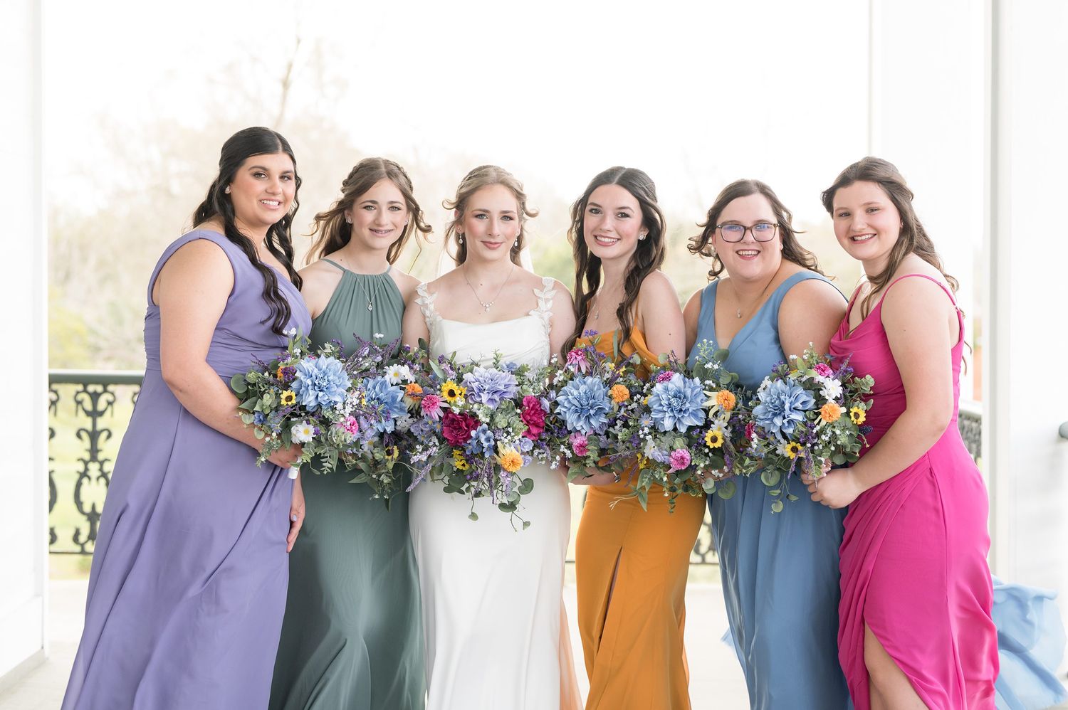 A group of bridesmaids in mismatched purple, green, and pink dresses holding colorful floral bouquets.