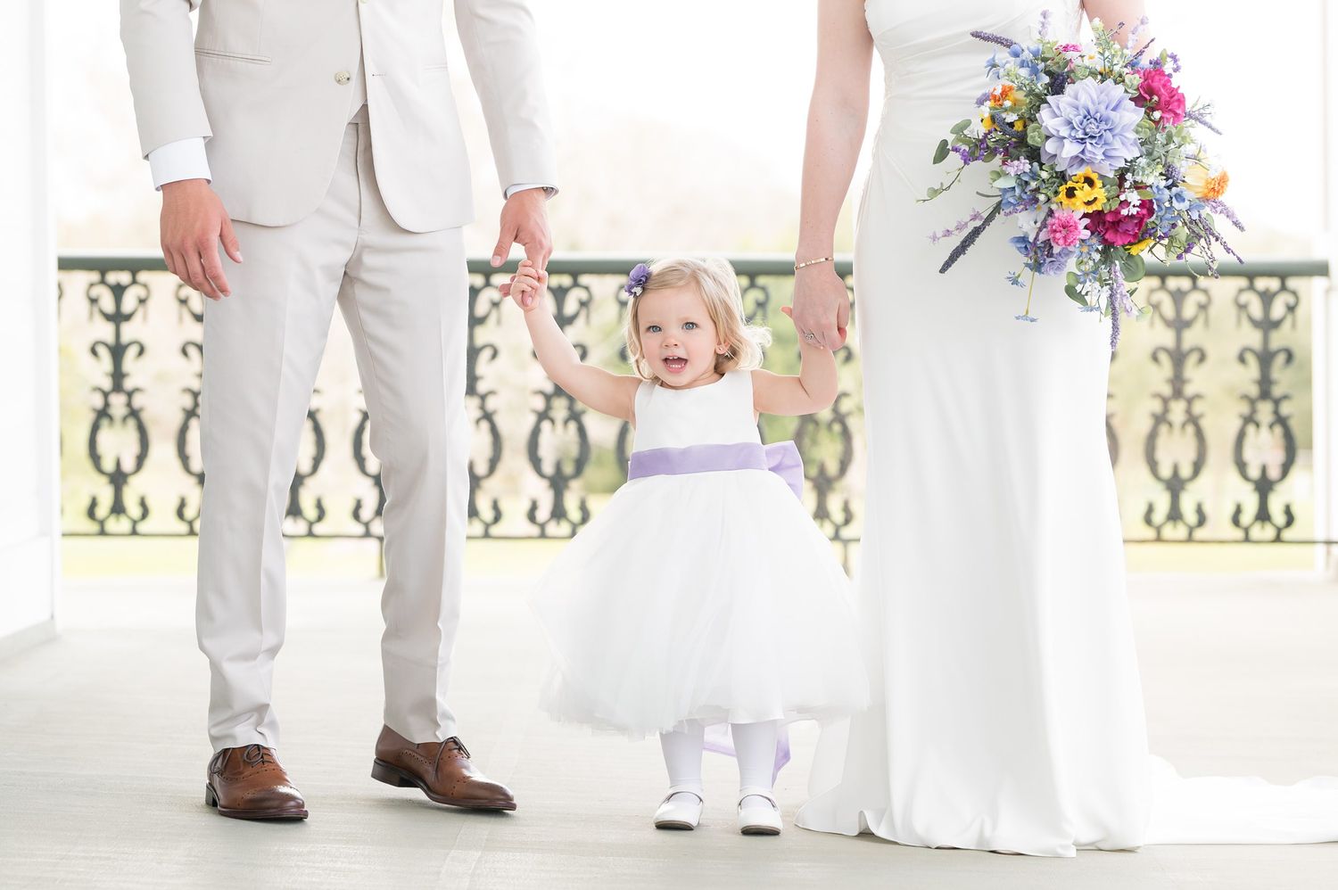 Wedding party wearing white and beige attire stands on an elegant porch with ornate railings and columns.