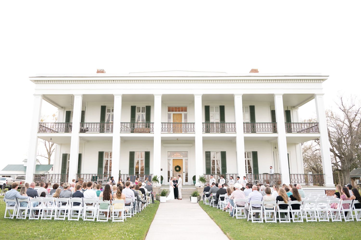 White folding chairs arranged in rows on a lawn in front of a grand mansion with tall columns for an outdoor ceremony.