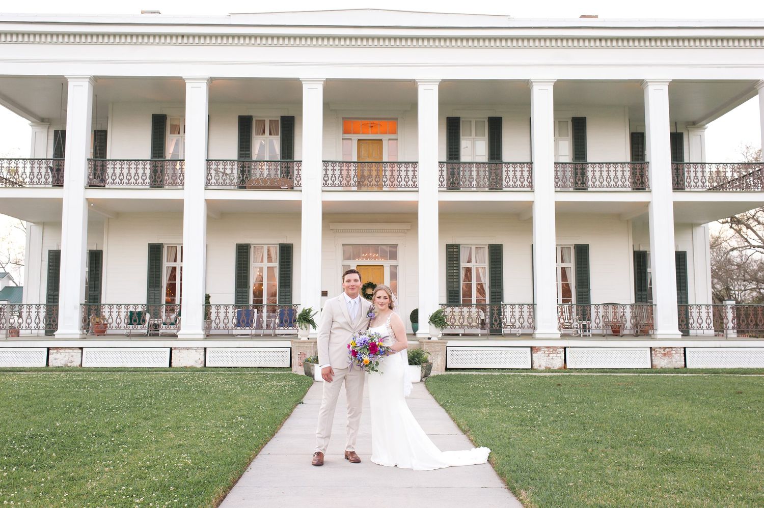 Elegant white antebellum mansion with two-story columns serves as backdrop for outdoor wedding ceremony.