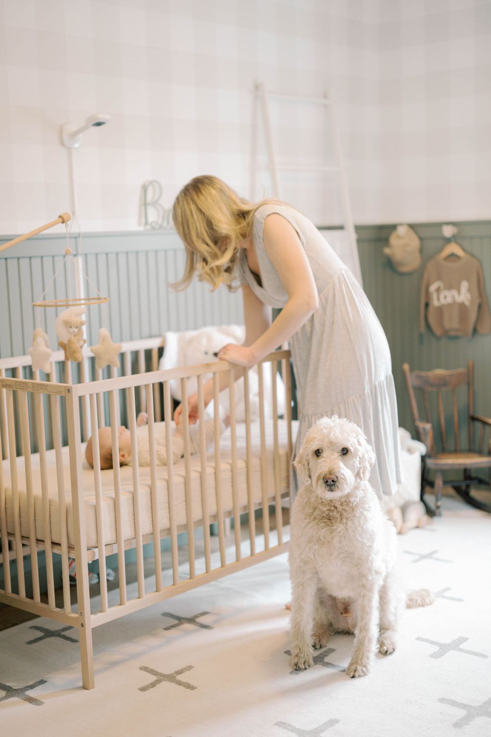 Blonde mother tending to newborn in light wood crib as white goldendoodle sits nearby in nursery.