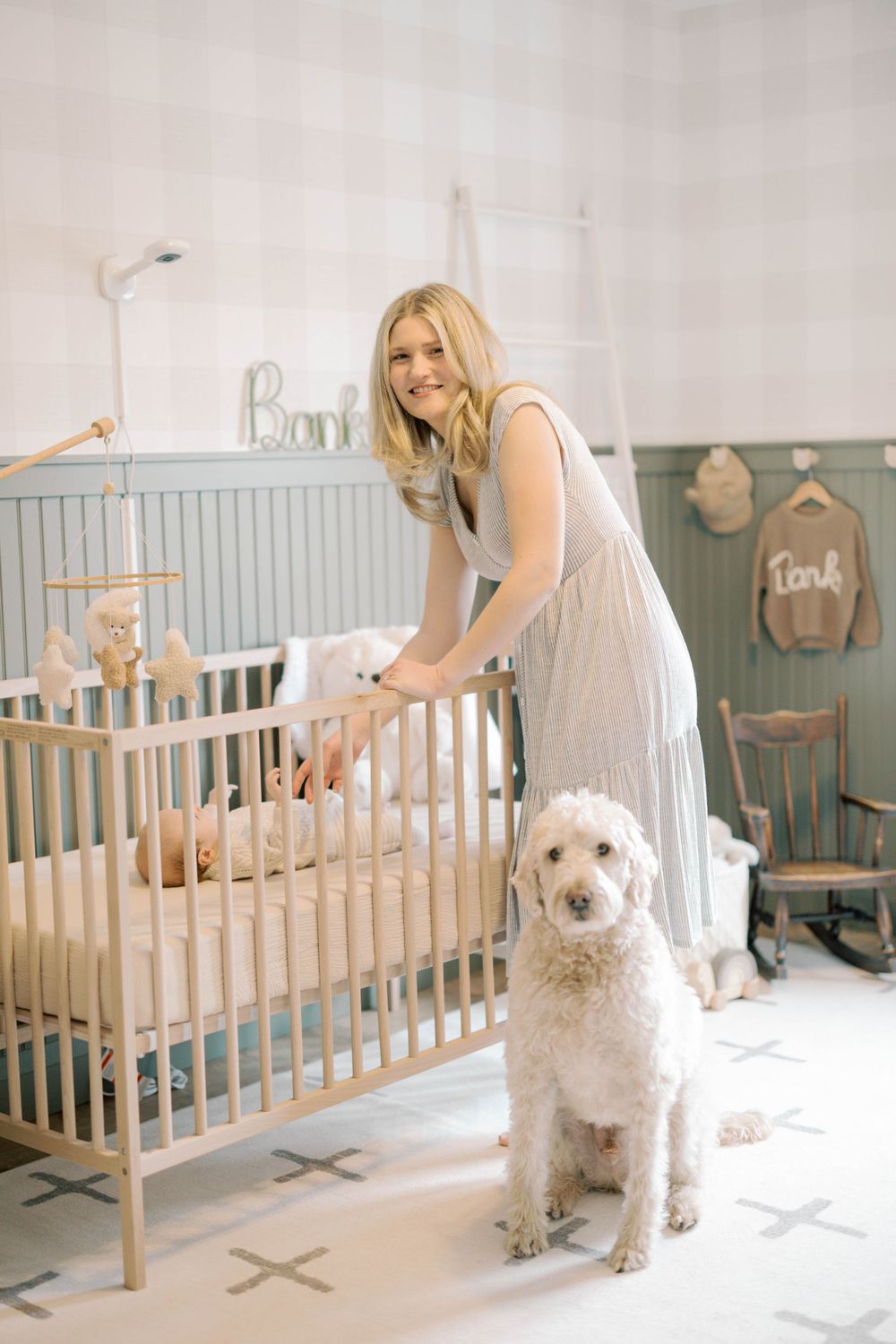 Smiling mother standing at light wood baby crib with white goldendoodle in a neutral sage nursery.