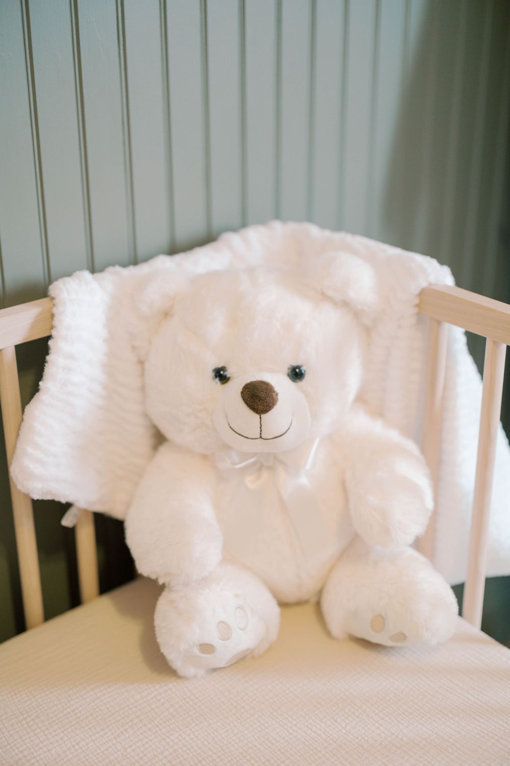 White plush teddy bear with bow tie sitting in a wooden baby crib with soft white blanket.