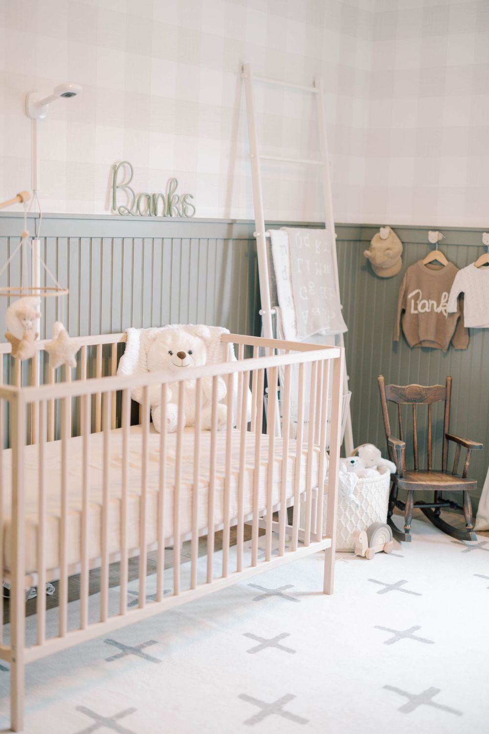 Neutral baby nursery with light wood crib, cross-pattern rug, sage green wainscoting, and personalized decor.