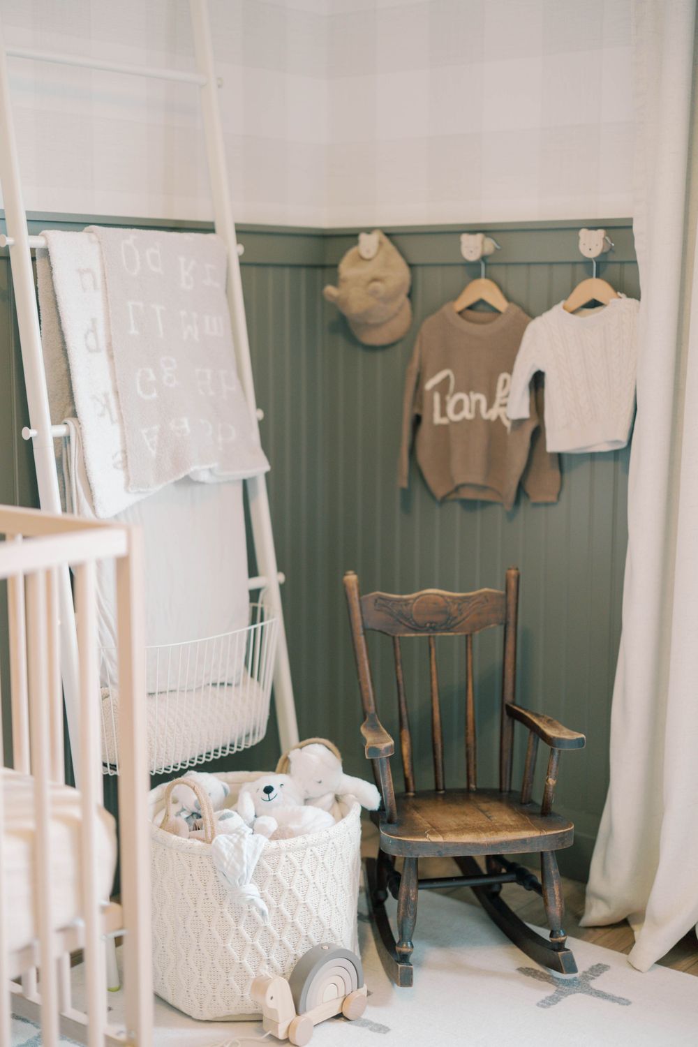 Sage green nursery corner with rocking chair, white knit basket, and personalized sweater hanging on wall hooks.