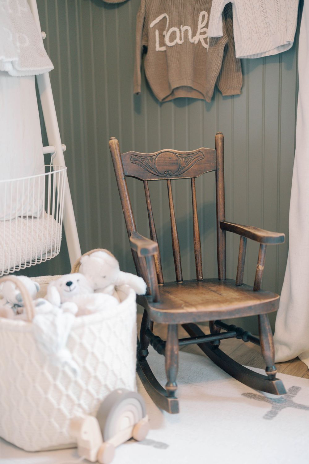 Vintage wooden rocking chair beside white knit toy basket in a sage green nursery with personalized sweater.