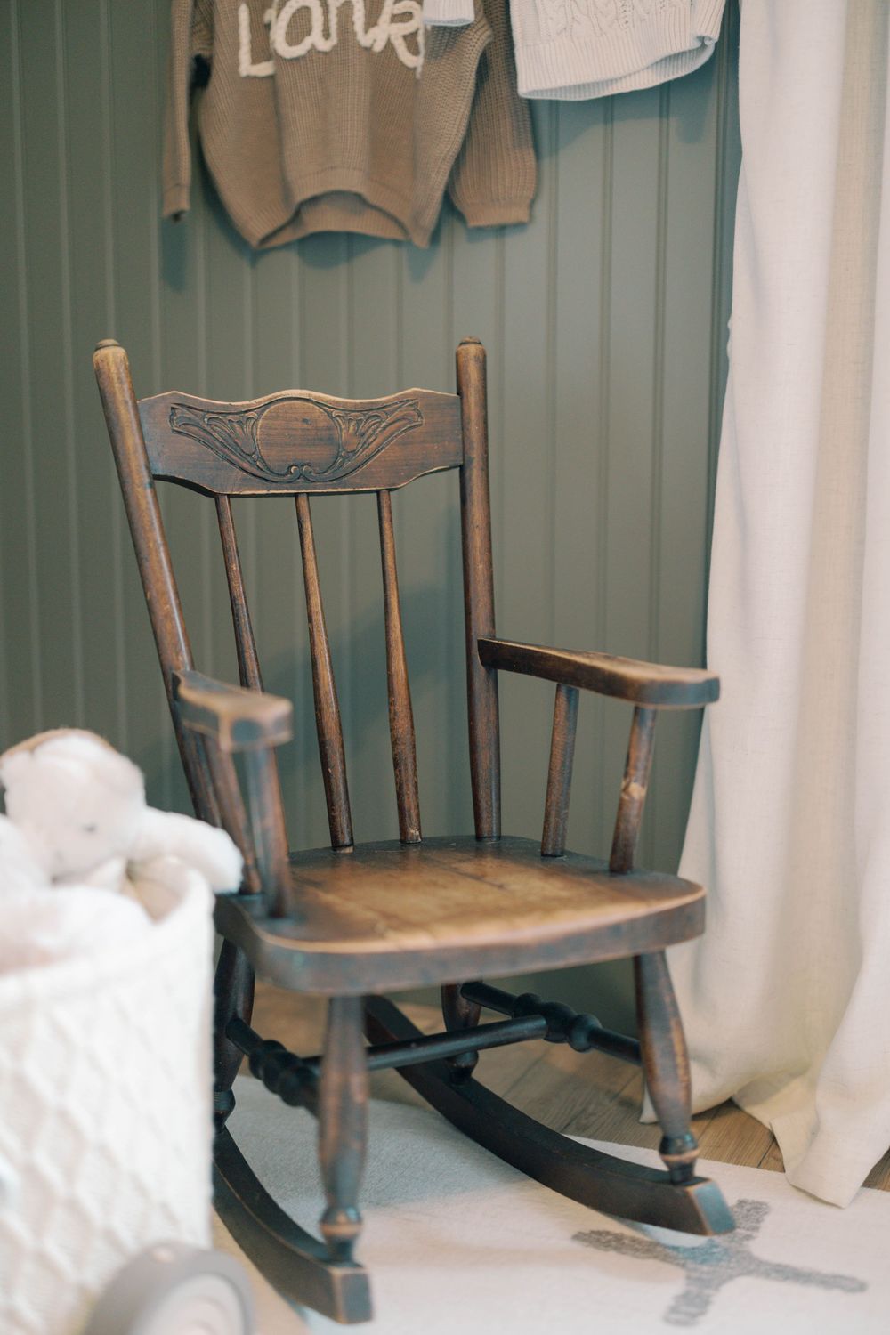 Vintage dark wood rocking chair in sage green nursery with hanging knit sweater and white wicker basket.