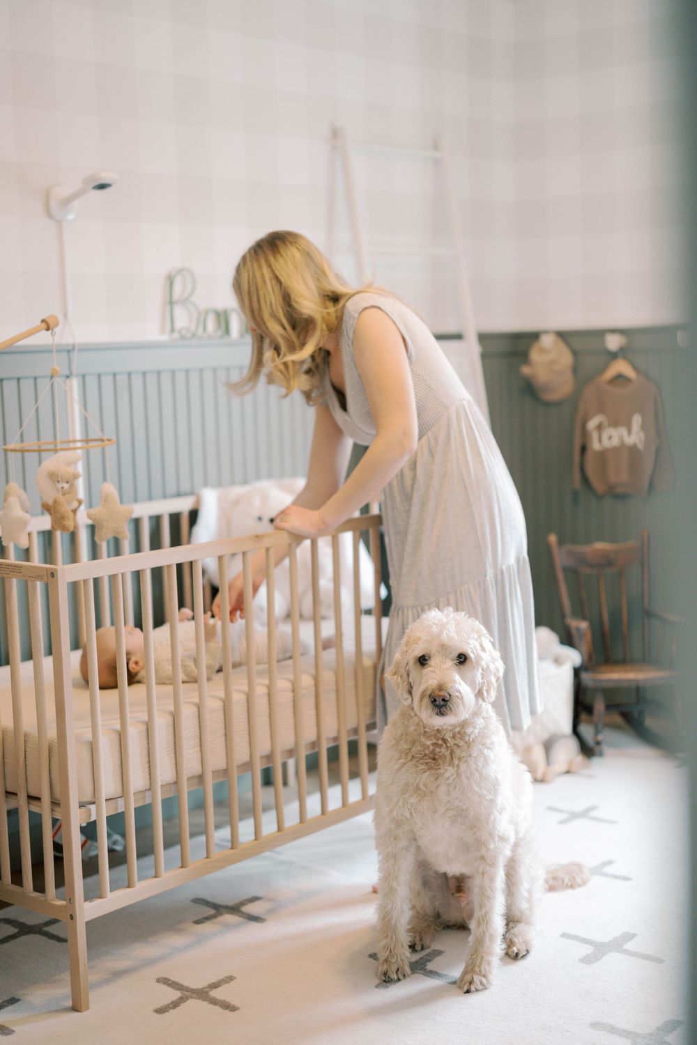 Blonde mother leans over wooden crib tending to newborn while white goldendoodle sits beside her in nursery.