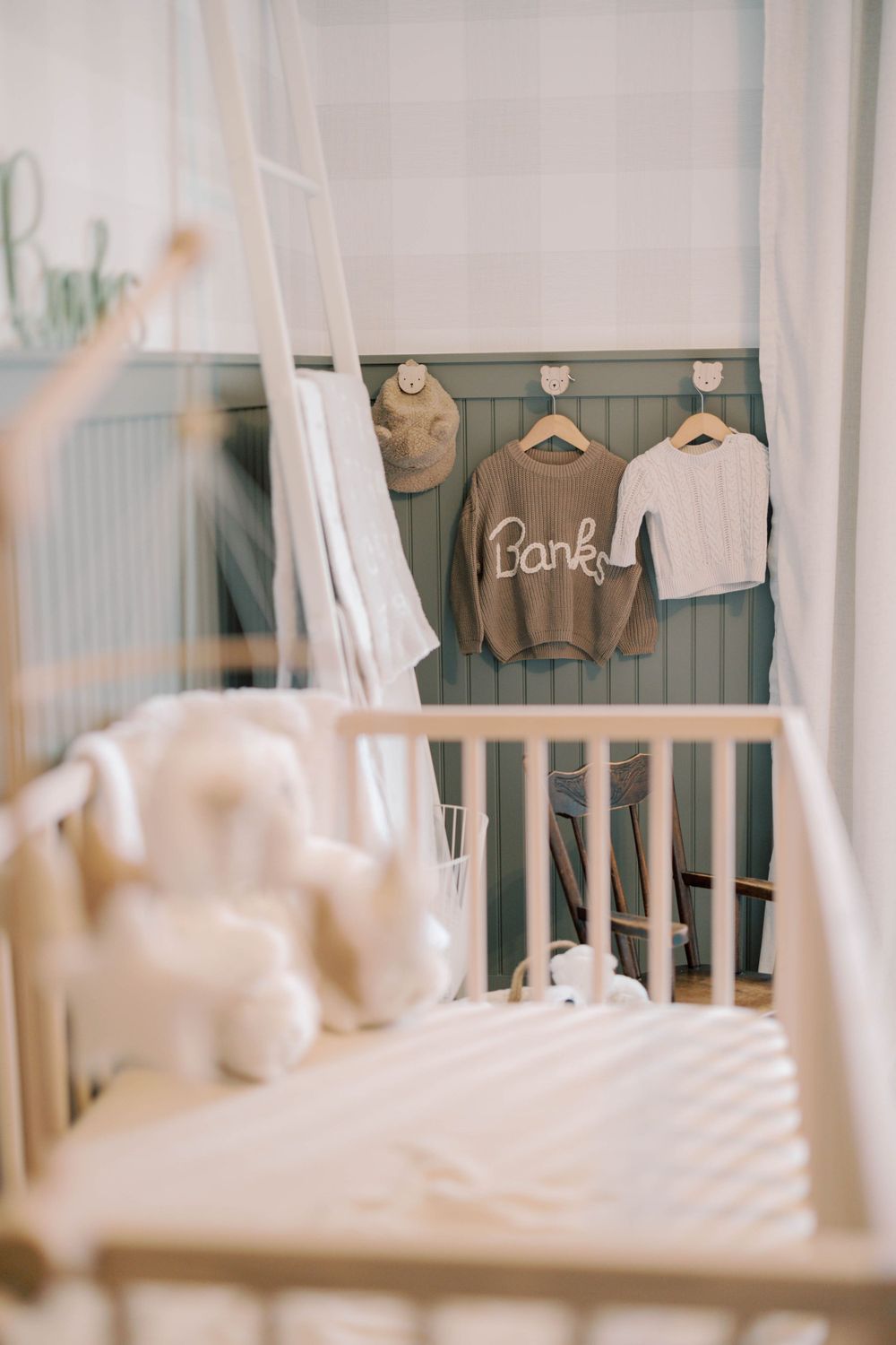Light wood baby crib with stuffed animals and personalized sweaters hanging on sage green beadboard wall.