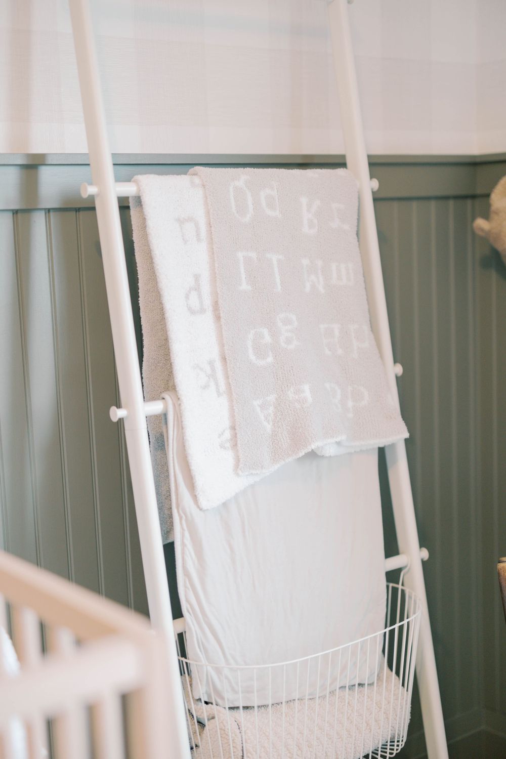 White alphabet baby blanket draped over a decorative ladder next to a wooden crib in a sage nursery.