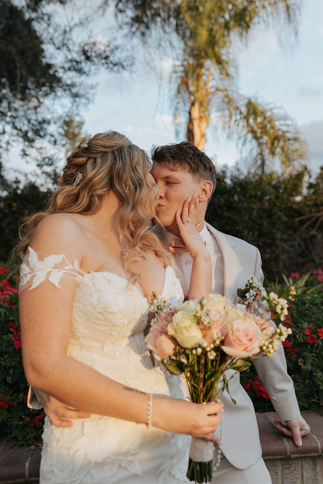 A romantic wedding moment captured outdoors with palm trees and a beautiful bouquet of pink and white roses.