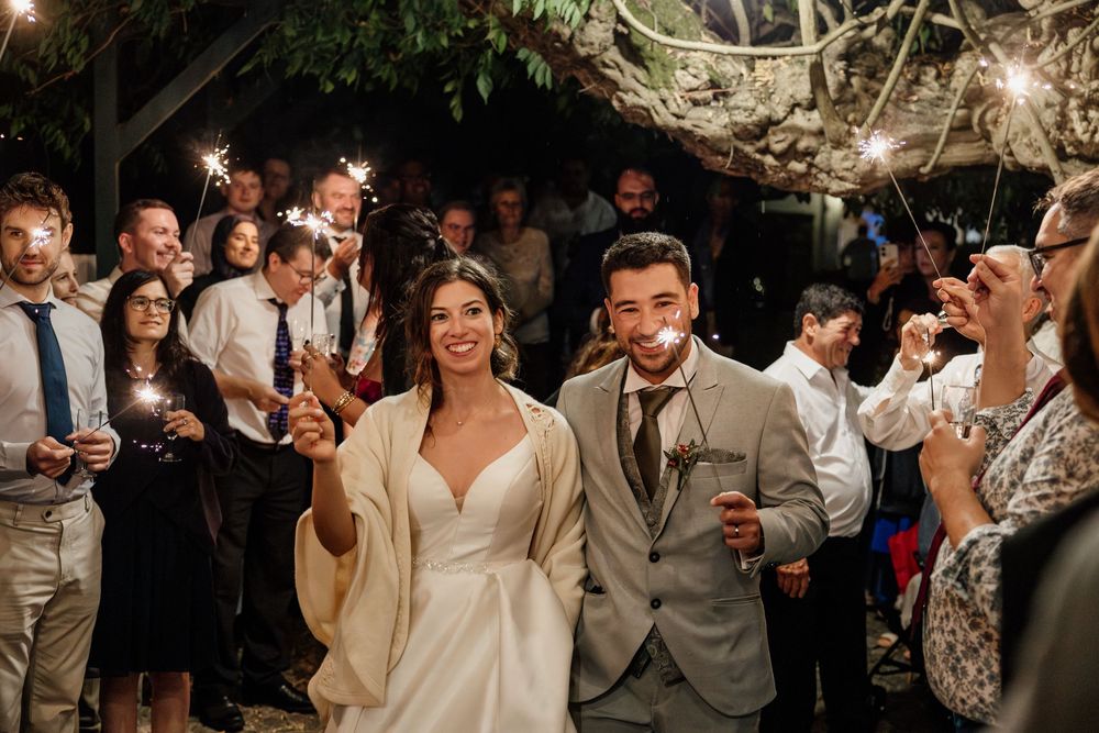 Bride and groom walk through sparkler exit surrounded by cheering wedding guests at an outdoor evening reception.