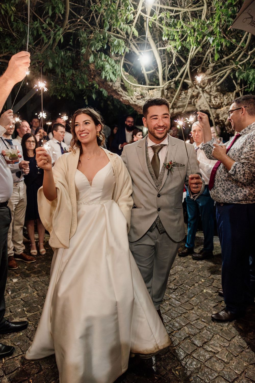 Bride and groom walk through sparkler exit surrounded by wedding guests at night outdoor ceremony.
