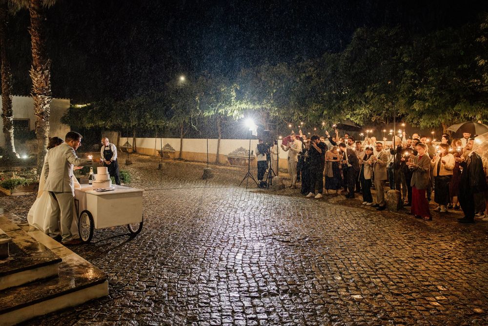 Couple cuts wedding cake outdoors at night while guests hold torches on cobblestone venue.