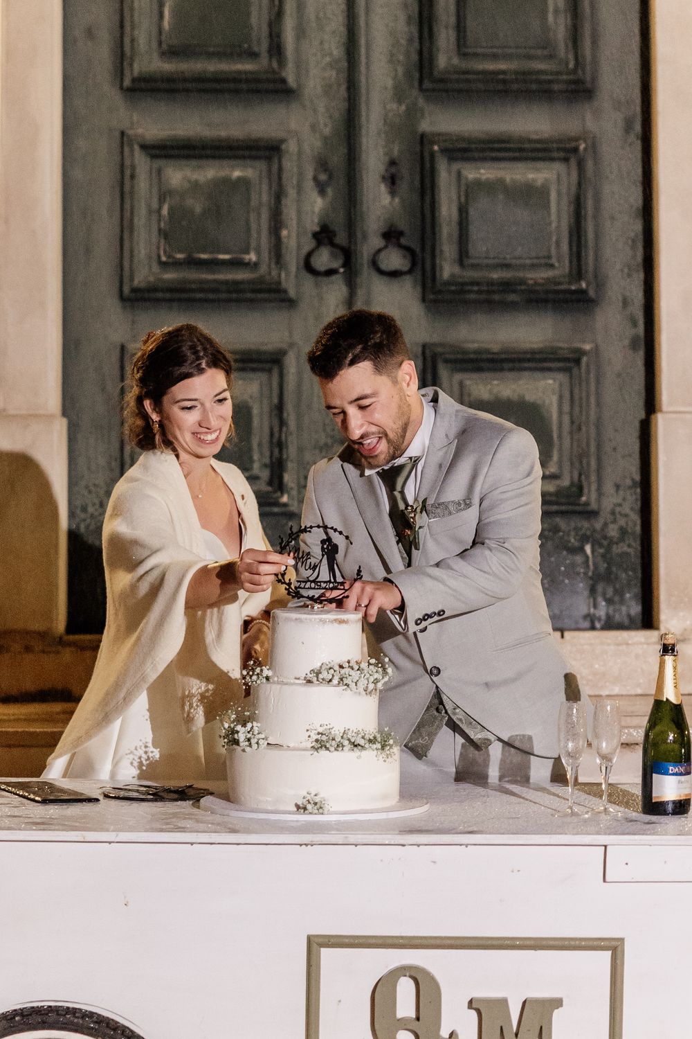 Bride and groom cutting white three-tier wedding cake with floral topper, champagne bottle nearby, elegant venue.