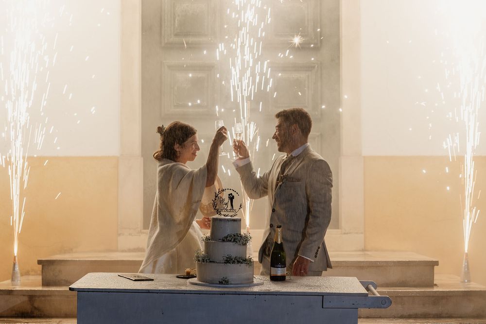 Bride and groom toasting champagne over wedding cake with sparkling fireworks display indoors at elegant venue.