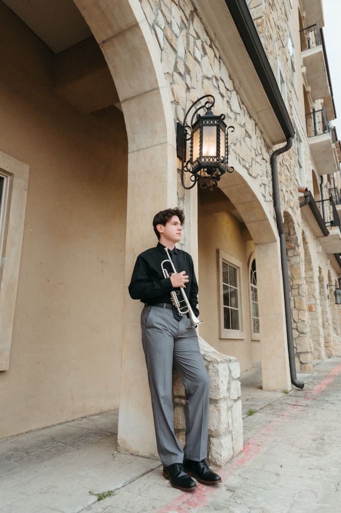 Young musician in black shirt and gray trousers holds trumpet while leaning against stone building with ornate lantern.