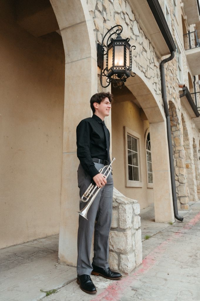 Young trumpet player in black shirt and gray pants poses by stone arch with decorative lantern.