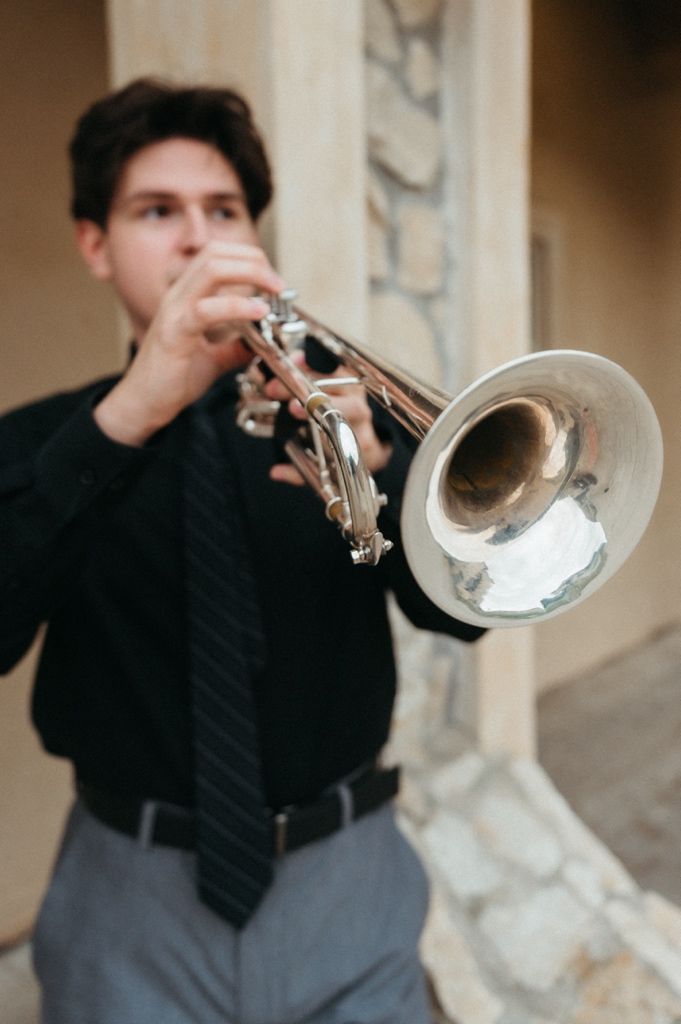 Young man in black shirt and tie playing a silver trumpet outdoors near a stone column.