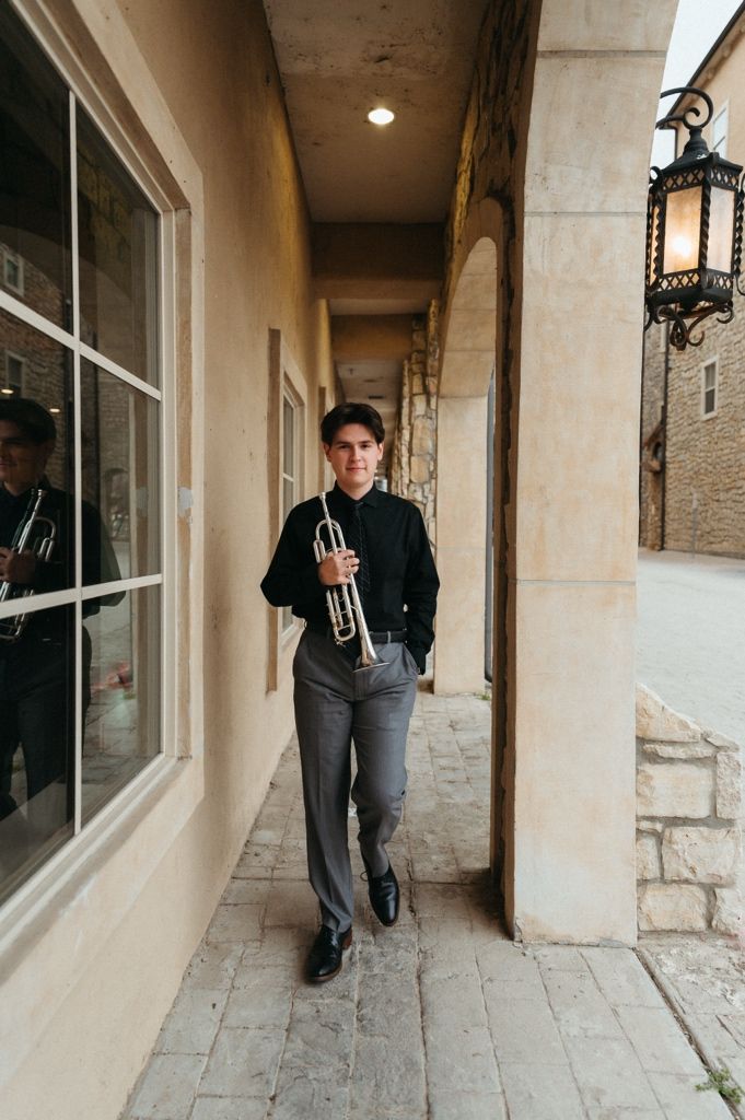 Young trumpet player in black shirt and gray pants walking along a covered outdoor corridor.