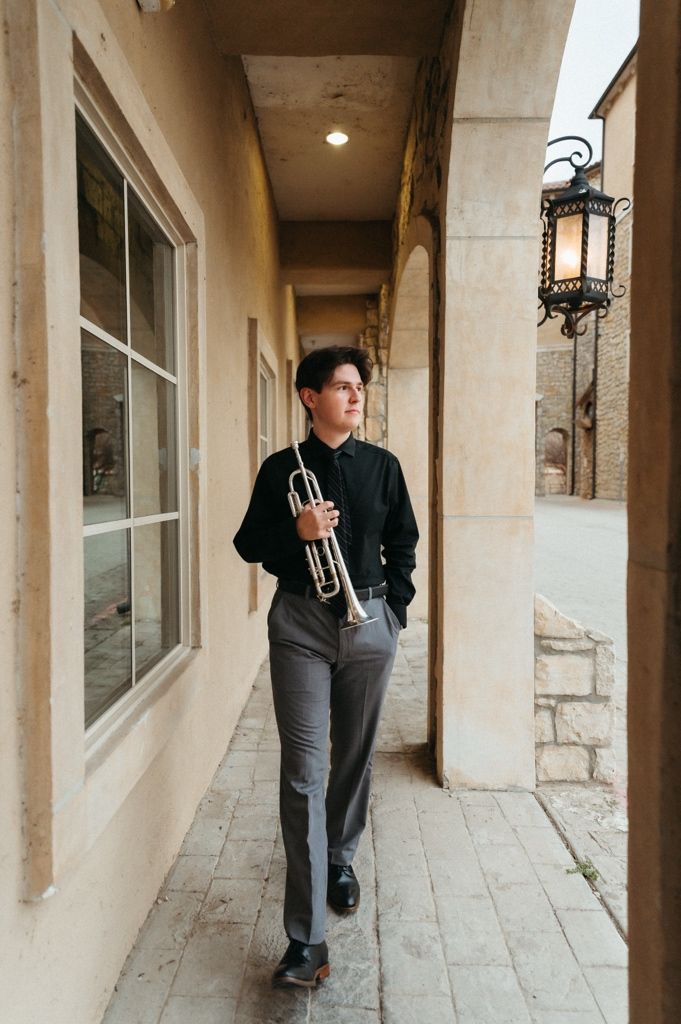 Trumpet musician in formal black shirt walks along stone building corridor holding his instrument.