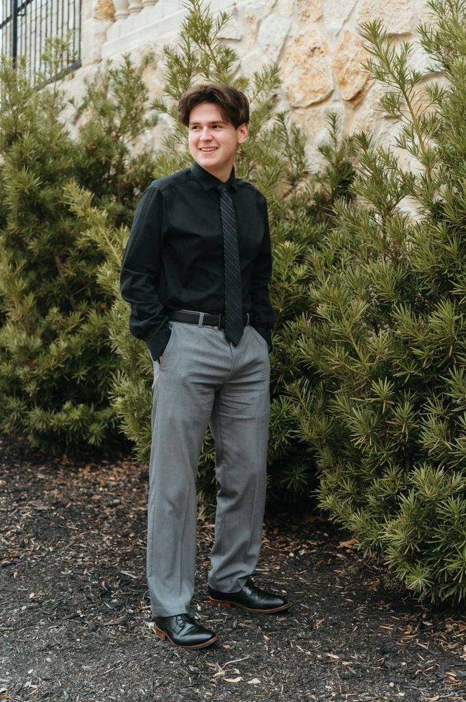 Young man in black dress shirt, gray slacks, and tie poses outdoors near evergreen shrubs and stone wall.