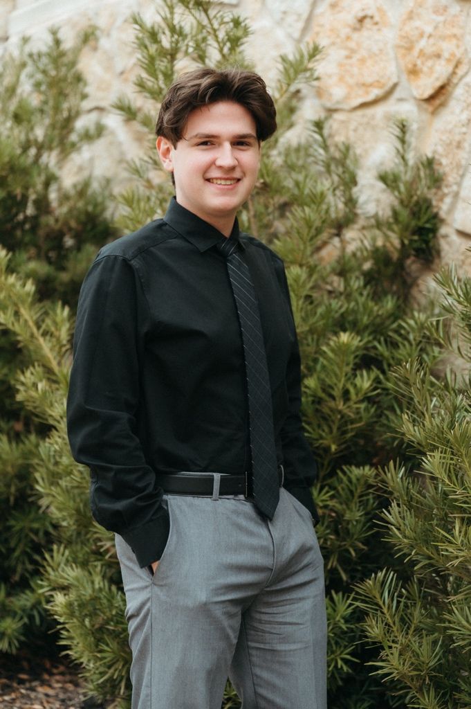 Young man in black dress shirt, gray tie, and gray slacks smiling outdoors near evergreen shrubs and stone wall.
